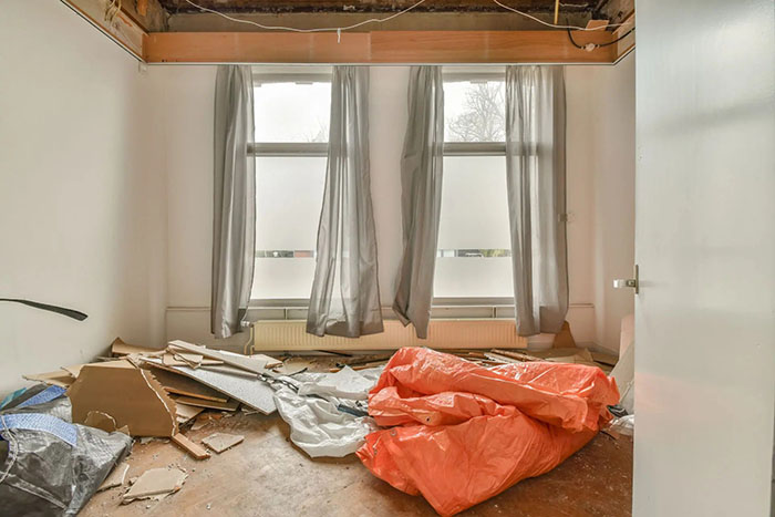 A room with a damaged ceiling, debris on the floor, and plastic sheets near the window.